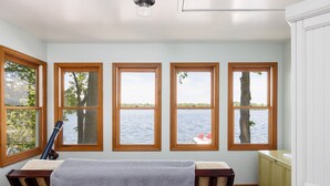 A room with a view of a lake, wooden-framed windows, a table with a blanket, and a chair.