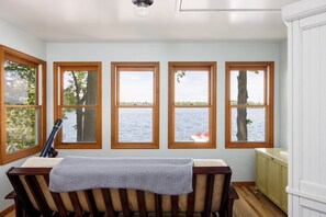 A room with a view of a lake, wooden-framed windows, a table with a blanket, and a chair. - Firefly Cottages on Bass Lake | Sandy Frontage, Dock, Kayaks, Near Downtown TC (Traverse City)