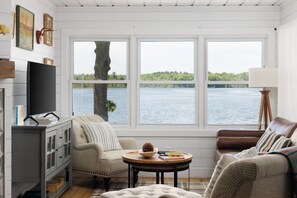 Ein Zimmer mit großem Fenster und Blick auf ein Gewässer, einem Fernseher, einem Kamin und einer gemütlichen Sitzecke.