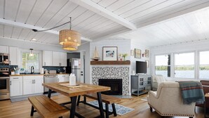 A modern kitchen with white cabinets, a dining table with benches, a fireplace, and a large window overlooking a body of water.
