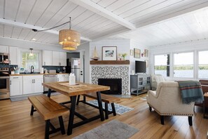 A modern kitchen with white cabinets, a dining table with benches, a fireplace, and a large window overlooking a body of water. - Firefly Cottages on Bass Lake | Sandy Frontage, Dock, Kayaks, Near Downtown TC (Traverse City)