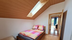 Ein Schlafzimmer mit schrägen Wänden, einem Bett mit farbenfroher Steppdecke, einer Bank mit gemustertem Kissen und Blick auf einen Balkon.