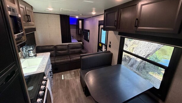 A compact RV interior with a kitchenette, seating area, and a window offering a view of the outdoors. - Rivers Edge RV Stay at Myakka Cove (Venice)
