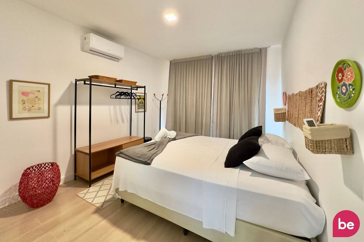 Um quarto com cama de solteiro, mesa de cabeceira, cadeira vermelha de fibra natural, ar condicionado fixado na parede e janela com cortinas.