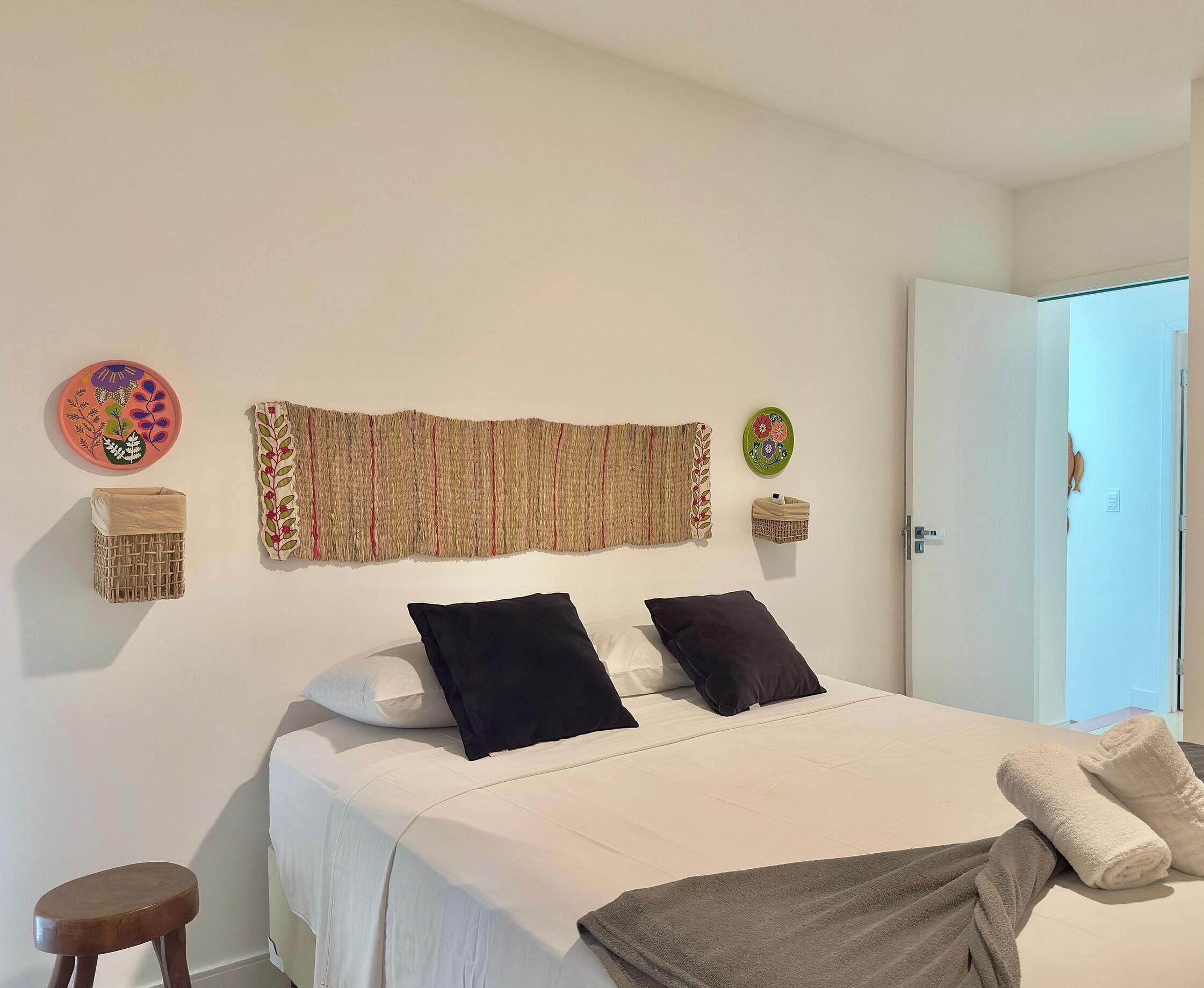 Una cama con ropa blanca, dos almohadas negras, una manta marrón y un taburete café.