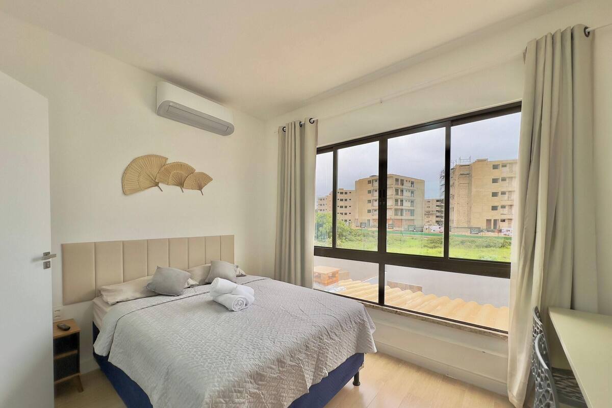 Quarto com cama, mesa de cabeceira, ar condicionado e uma janela grande com cortinas.