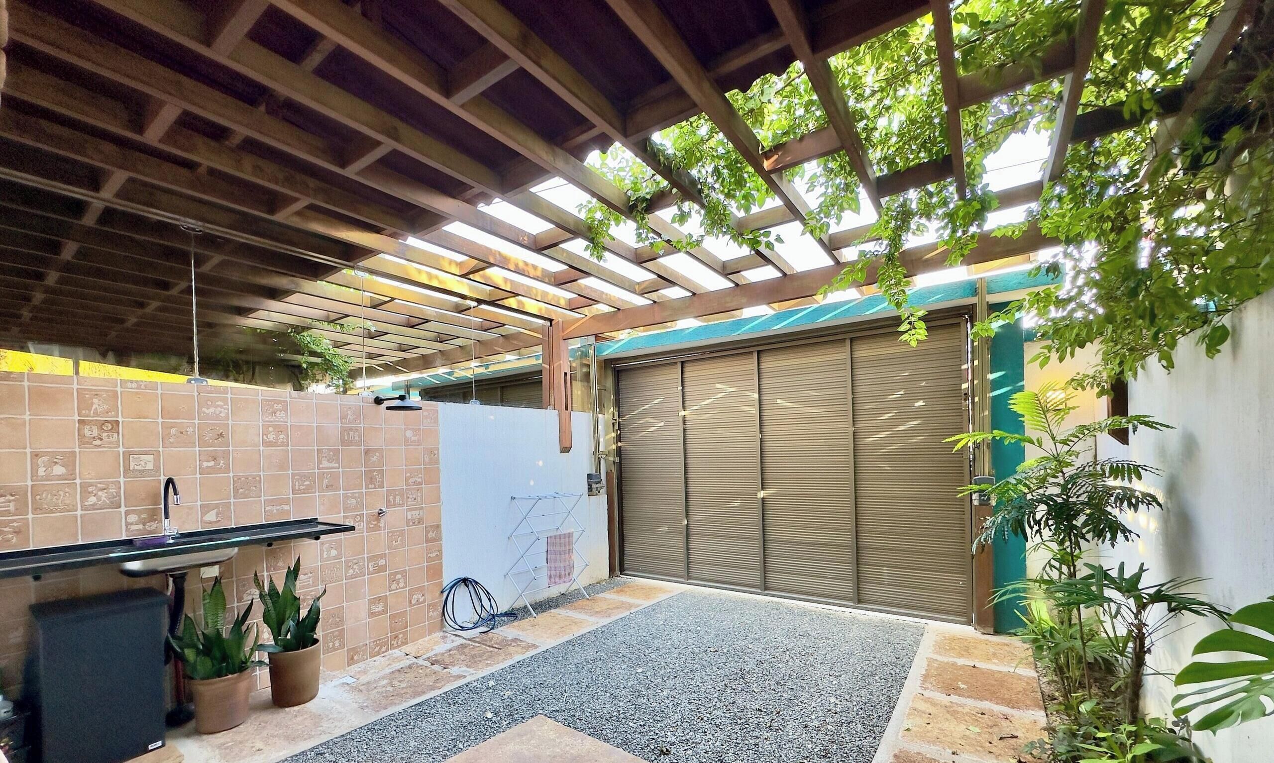 Un patio couvert avec un sol carrelé, un banc en métal, des plantes en pot et une porte à enroulement fermée.