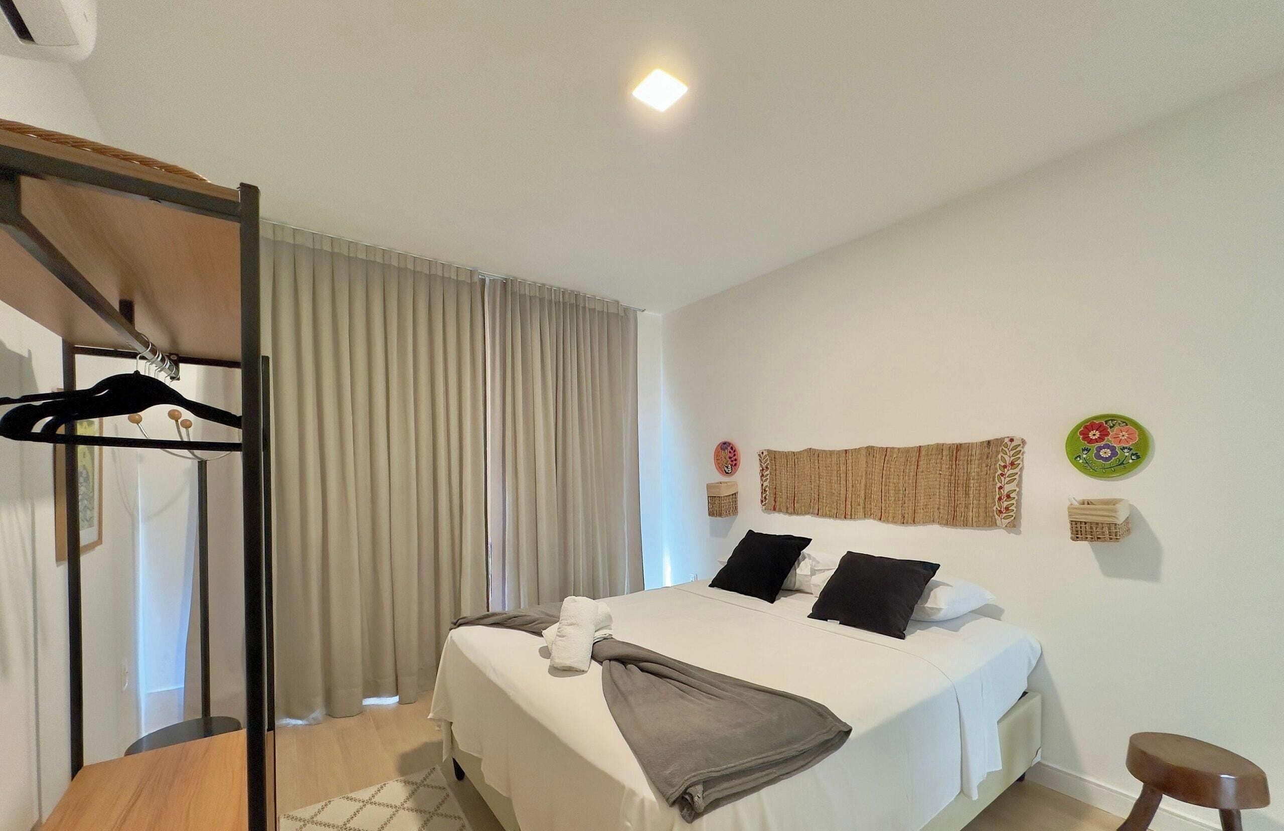 A hotel room with a bed, a wooden chair, a coat rack, and a window with curtains.