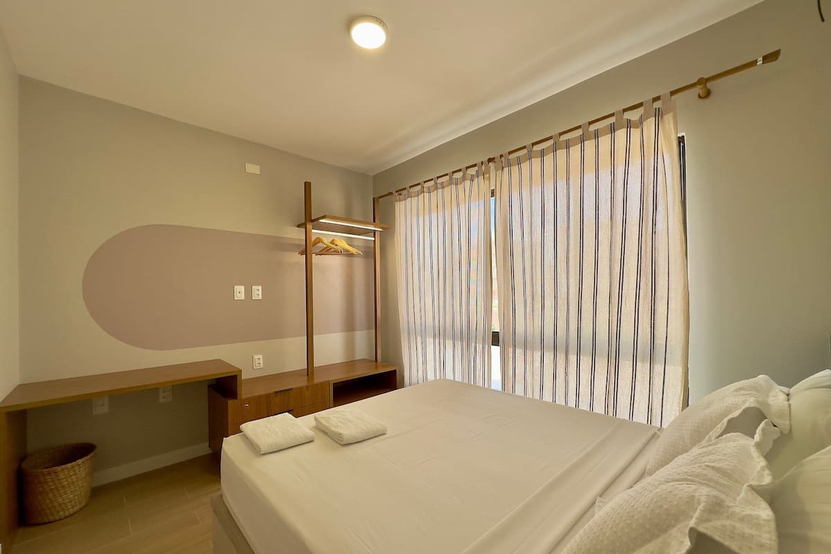 Quarto de hotel com cama, um banco de madeira, cabides suspensos e janela com cortinas listradas.