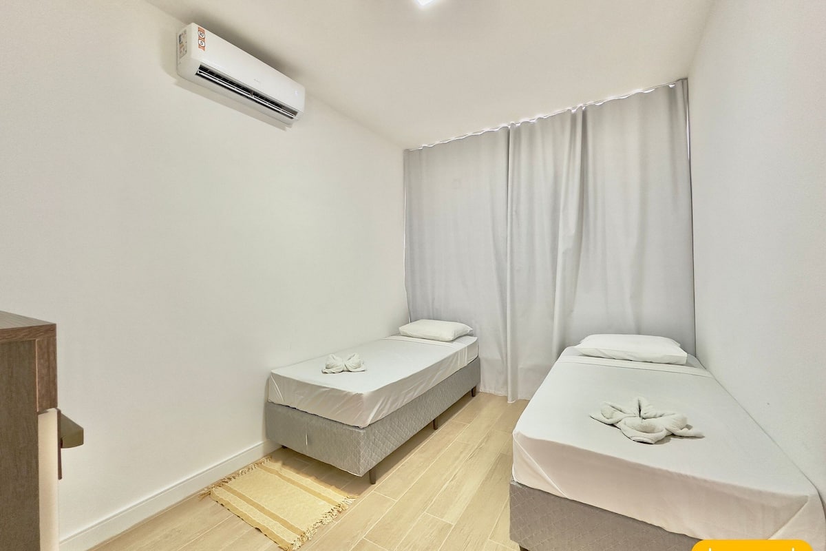 Quarto pequeno e limpo, com duas camas, uma mesa de cabeceira de madeira, um tapete bege e um ar condicionado branco.