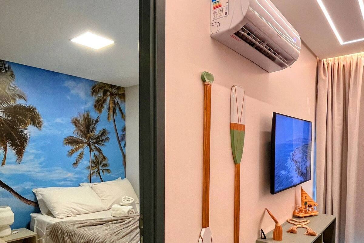 A hotel room with a bed, a TV, an air conditioner, and a wall with a palm tree mural.