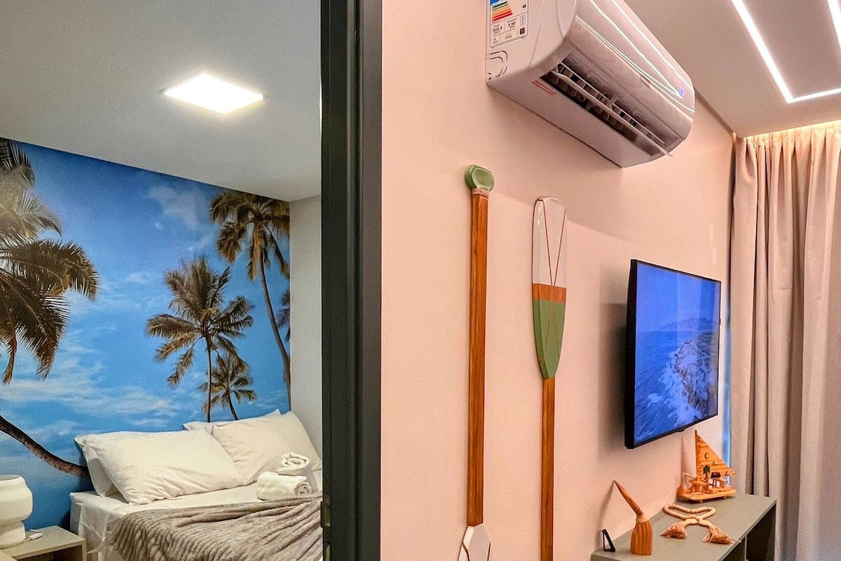 A hotel room with a bed, a TV, an air conditioner, and a wall with a palm tree mural.