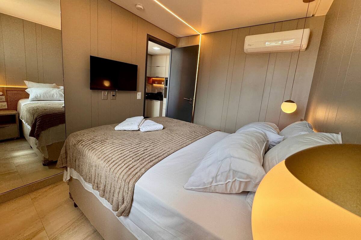 A hotel room with a bed, a TV, a mirror, and an air conditioning unit.