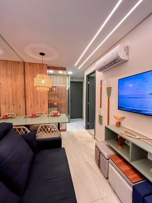 A modern living room with a sofa, TV, and dining area. - Vila Porto Beira Mar - VSH (Ipojuca)