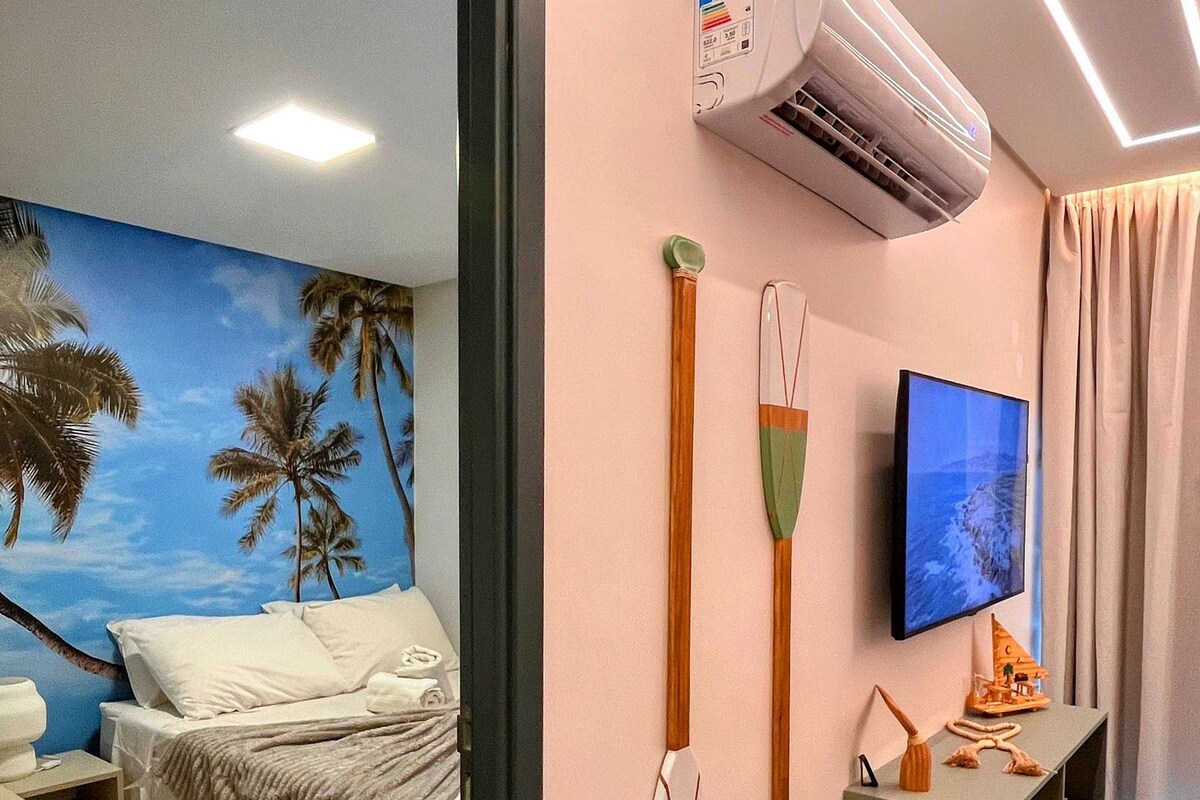 A hotel room with a bed, a TV, an air conditioner, and a wall with a palm tree mural.