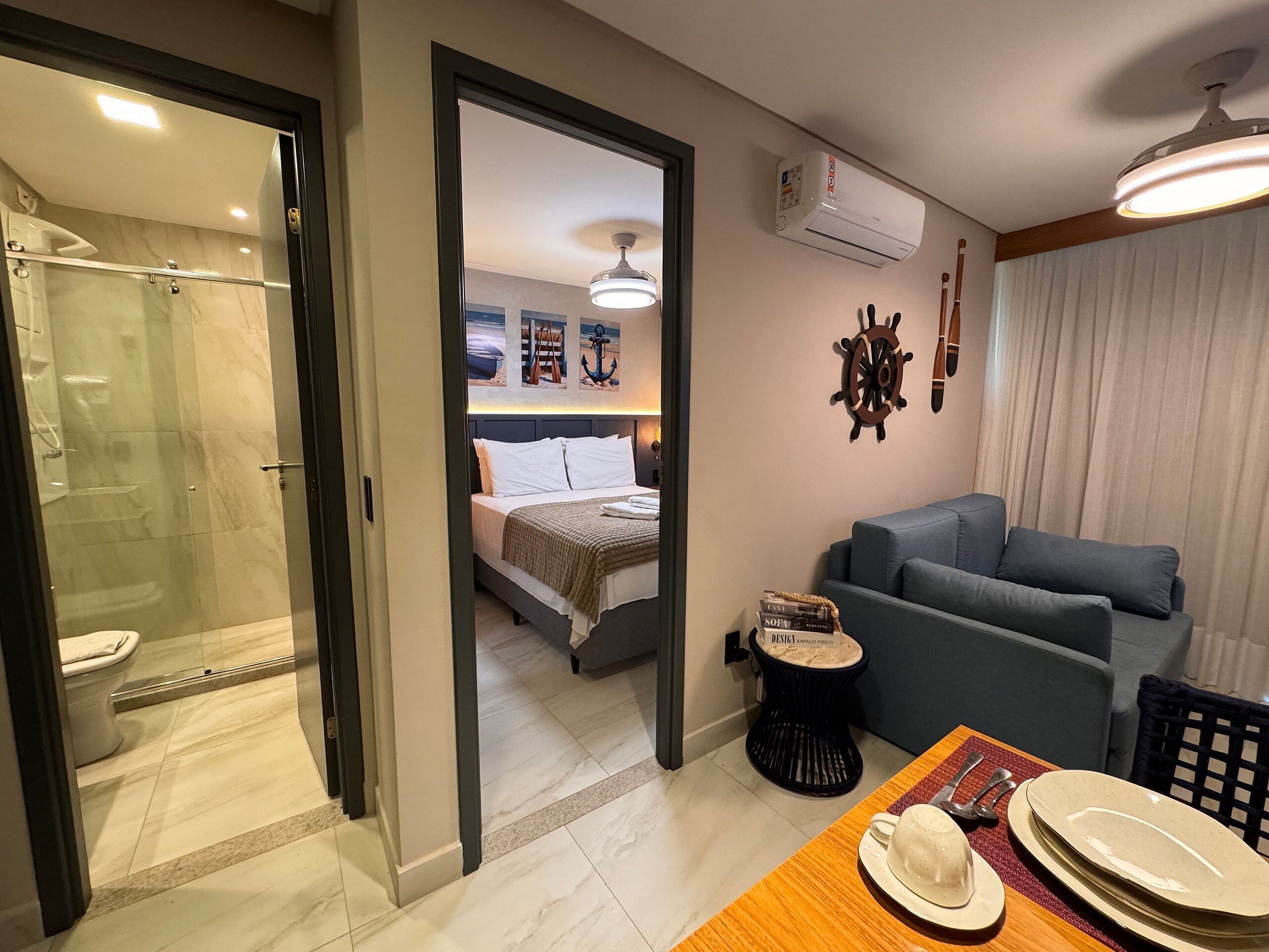 Habitación de hotel con ducha envuelta en vidrio, cama con ropa blanca, sofá gris, una pequeña mesa redonda con un plato y un sombrero, y aire acondicionado instalado en la pared.