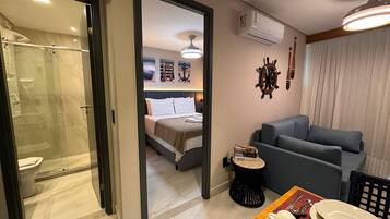 Habitación de hotel con ducha envuelta en vidrio, cama con ropa blanca, sofá gris, una pequeña mesa redonda con un plato y un sombrero, y aire acondicionado instalado en la pared.
