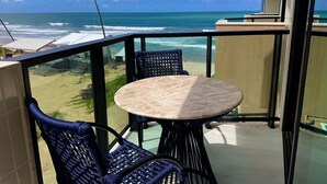 A balcony with a round table and two chairs, overlooking a beach with the ocean and a palm tree. - Vila Porto Beira Mar - VSH (Ipojuca)