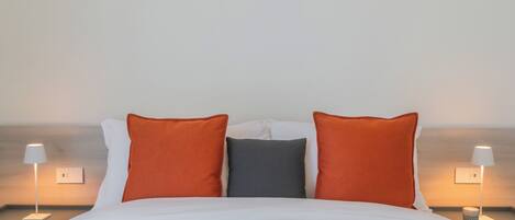 A neatly made bed with white and grey bedding, two orange pillows, and a grey pillow. There are two bedside lamps on either side of the bed.