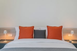 Una cama bien hecha con ropa de cama blanca y gris, dos almohadas naranjas y una almohada gris. Hay dos lámparas de noche a cada lado de la cama.