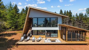 A modern two-story house with large windows, a deck with lounge chairs, and a forested backdrop.