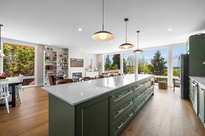 A modern kitchen with a large island, pendant lighting, and a view of trees through large windows.