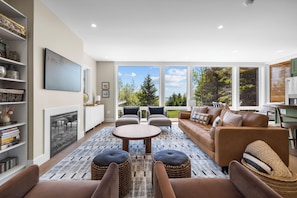 A living room with a fireplace, sofa, chairs, and a large window with a view of trees.