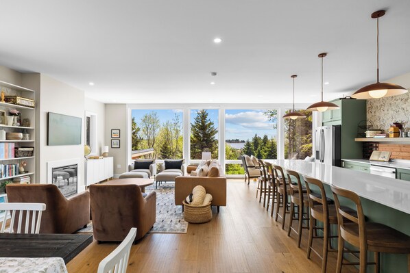 A modern kitchen with a large island, dining area with bar stools, and a living room with sofas and a fireplace.
