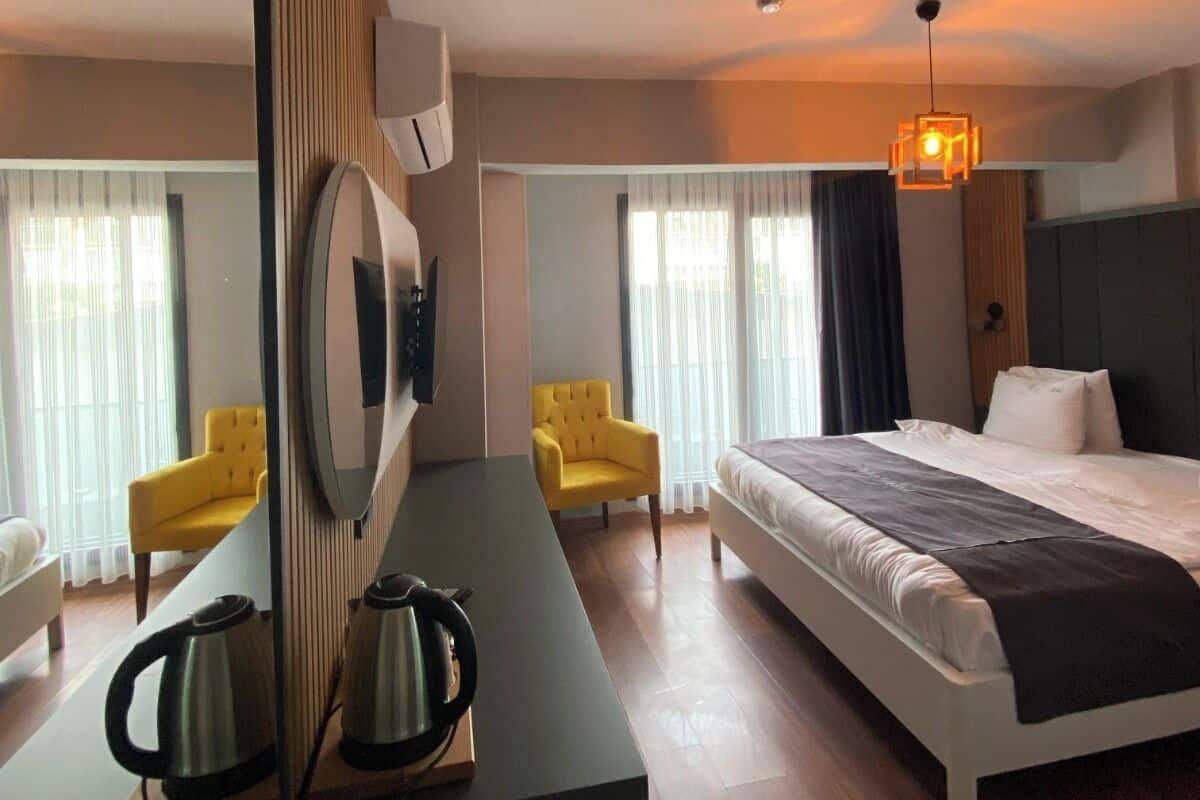 A hotel room with a bed, two chairs, a desk with a kettle, a mirror, and a hanging light fixture.
