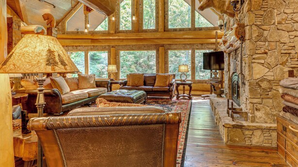 L’intérieur d’une cabane en rondins, avec une cheminée en pierre, des meubles en cuir et de grandes fenêtres.