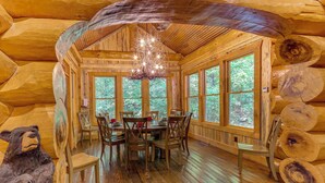 L’intérieur d’une cabane en rondins, avec une table à manger, des chaises, un lustre et de grandes fenêtres.
