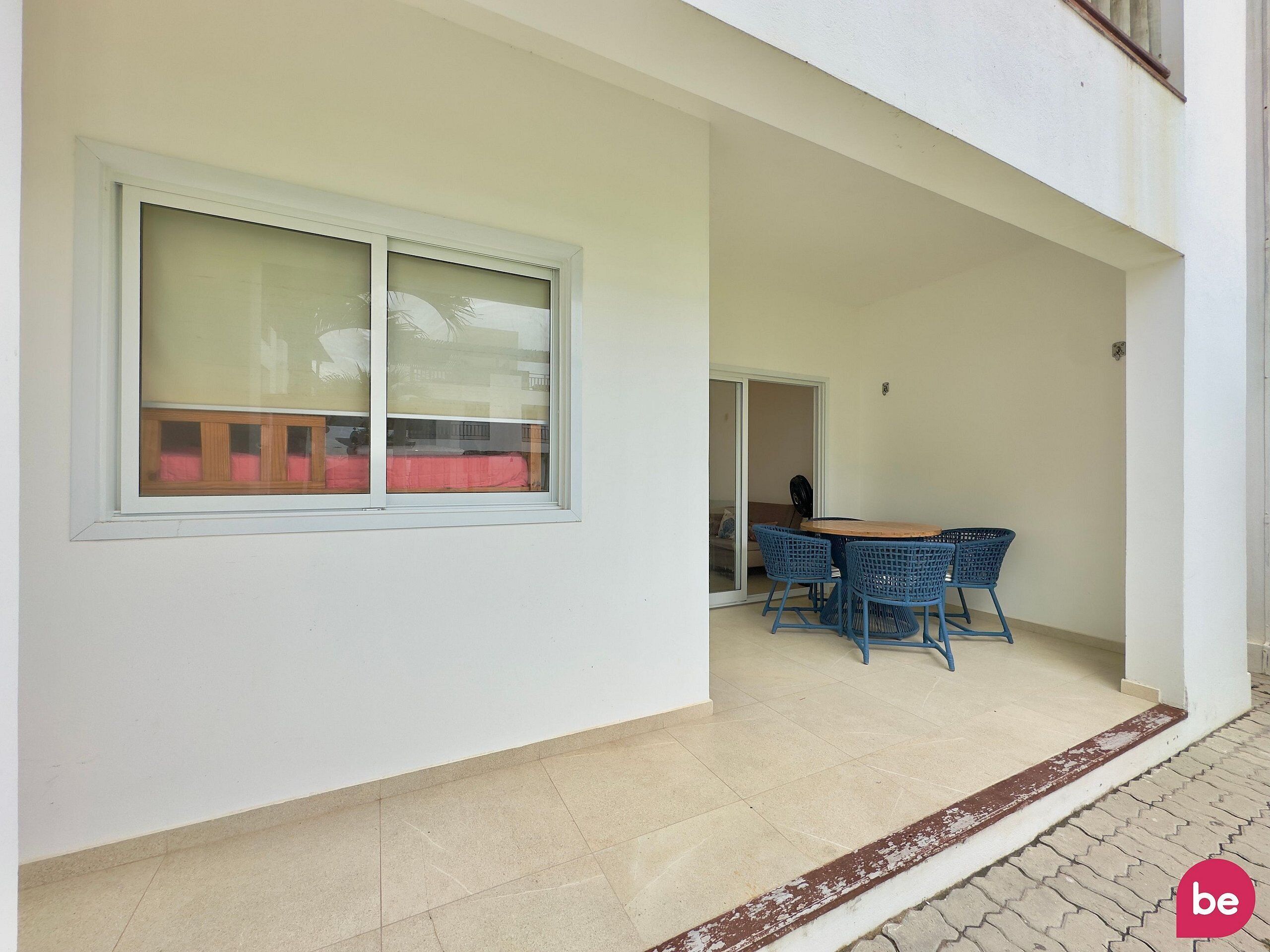 A covered outdoor seating area with a table and chairs, a sliding glass door leading to an interior space, and a tiled floor.