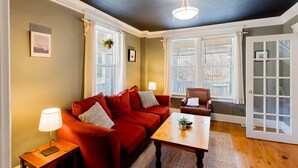 Ein Zimmer mit einem roten Sofa, einem Holztisch, einer Lampe und einem Sessel. Zwei Fenster mit weißen Rahmen und eine Glastür sind vorhanden.
