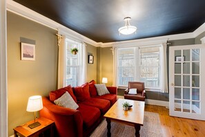 A room with a red sofa, a wooden coffee table, a lamp, and a chair. There are two windows with white frames and a door with glass panels. - The Porch Light - 5 Bedroom Retreat with music room. (Hope River)