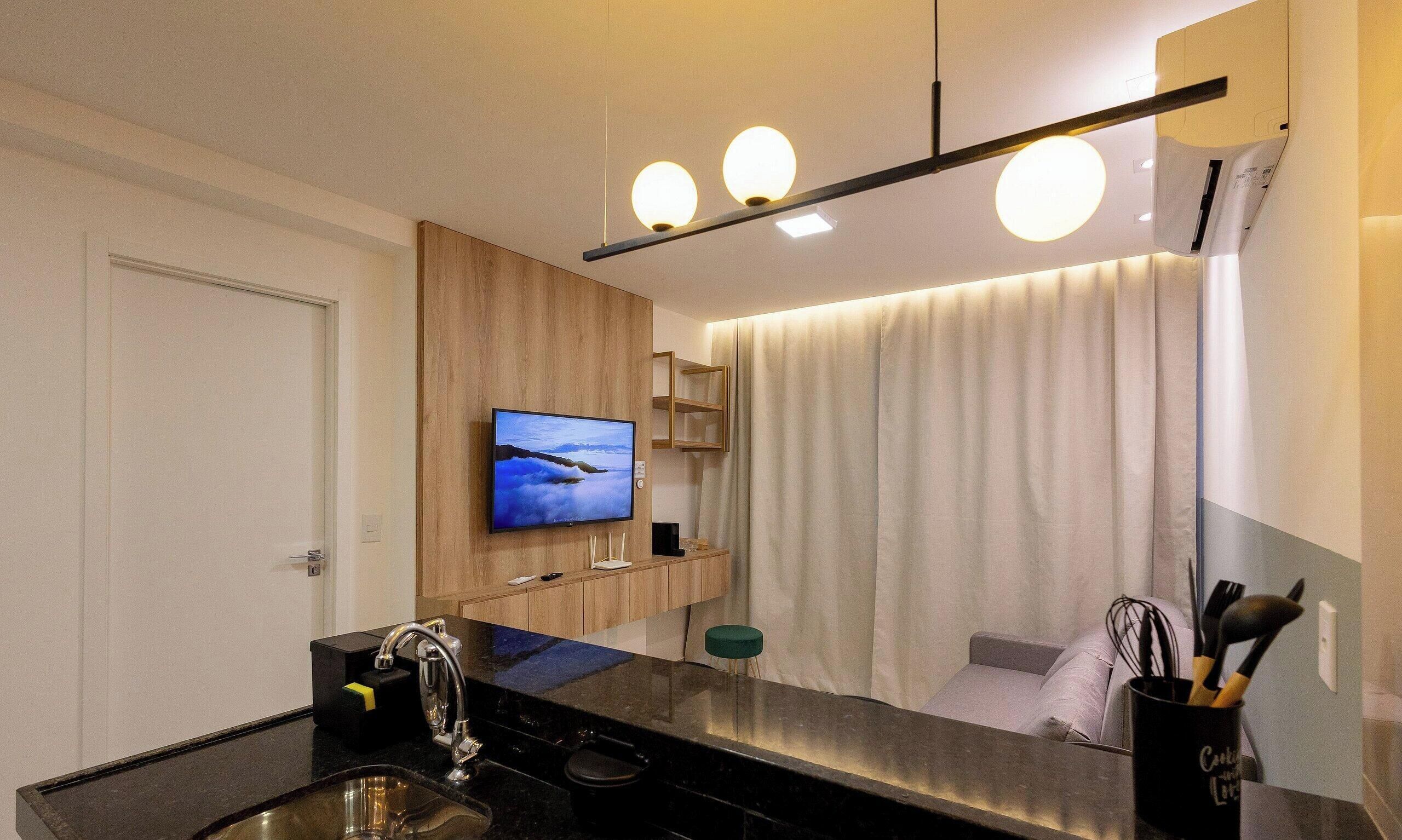 A modern kitchen with a flat-screen TV, wooden cabinetry, a black countertop, a sink, and a mirror.