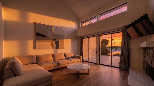 A modern living room with a sofa, a coffee table, and a large window with a view of the sunset. - Quiet Lakefront Home with Boat, Hot Tub, Game Room, Great Views, Sleeps 10 (Gilbert)