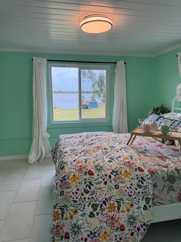 A bedroom with a large window, a bed with a floral bedspread, a bedside table with a clock, and a view of a lake.