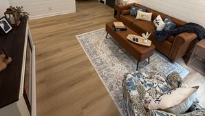 Ein Wohnzimmer mit braunem Sofa, einem gemusterten Hocker und einem Essbereich mit Tisch und Stühlen.