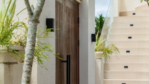 Un sendero conduce a un edificio con puerta de madera, grandes jarrones decorativos y una palmera.