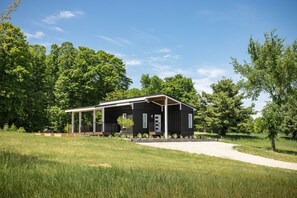 A modern black cabin with a covered porch, surrounded by a grassy field and trees. - Cargo Cozy - countryside cottage with hot tub (Suttons Bay)