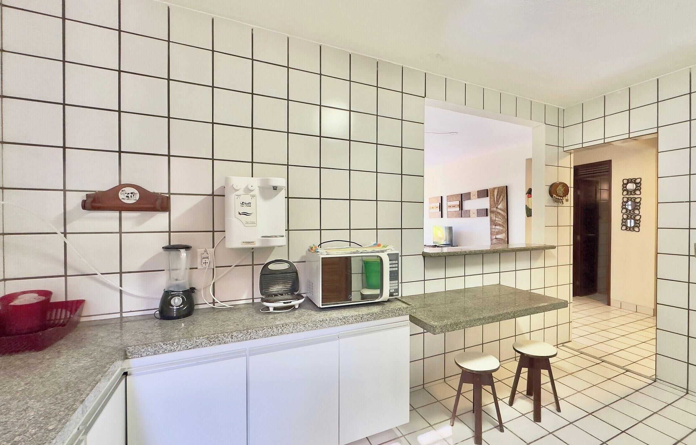 Una cocina con azulejos blancos, una licuadora, un microondas y una pequeña barra con taburetes.