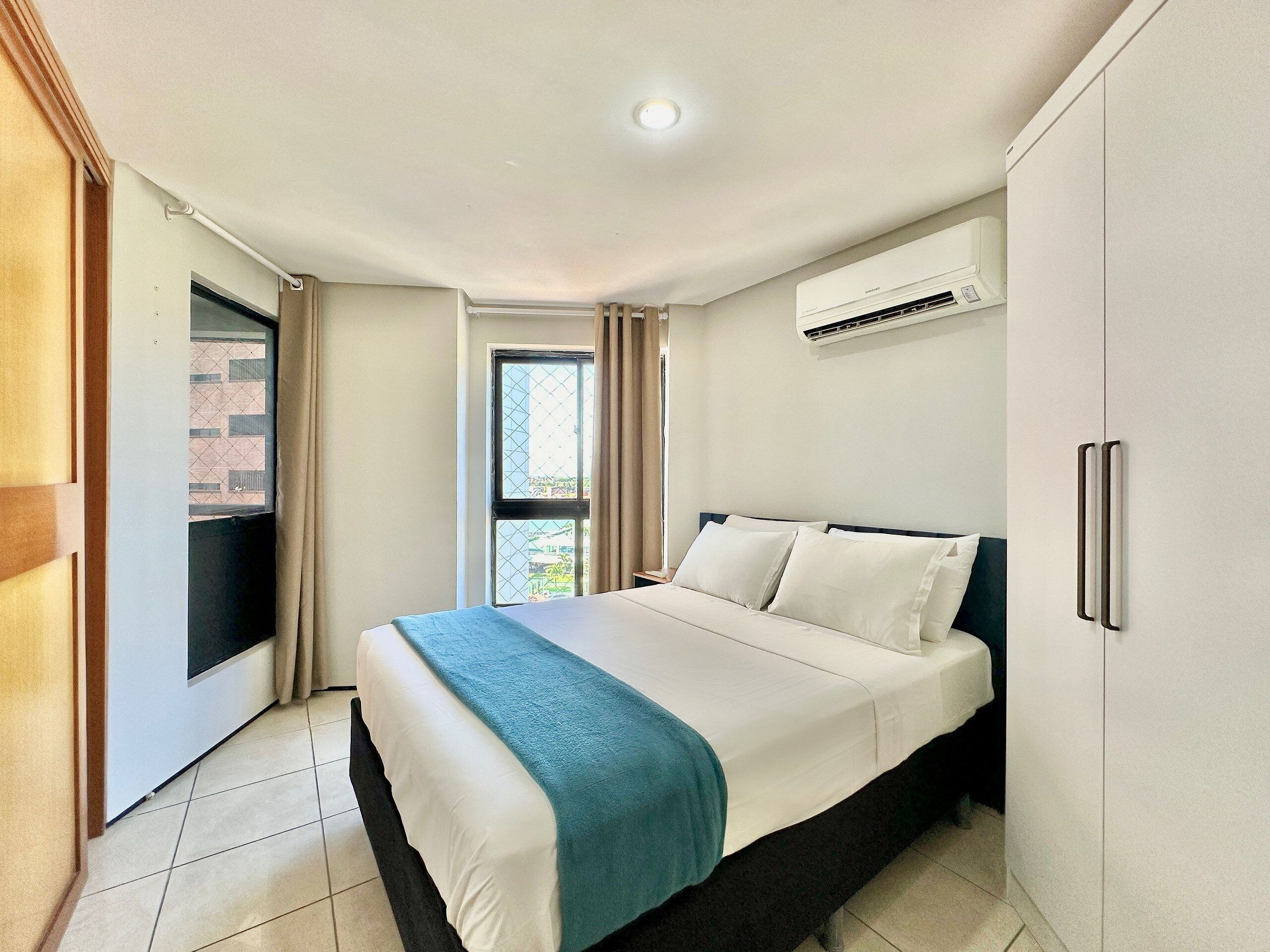 A hotel room with a bed, an air conditioning unit, a window with curtains, and a door leading to an outdoor area.