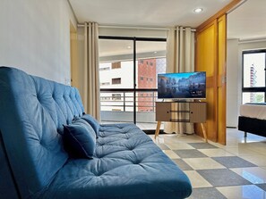 A modern living room with a blue sofa, a television on a stand, and a view of a building through a glass door. - Apart-hotel Iate Plaza, Vista Completa da Orla de Fortaleza (Fortaleza)