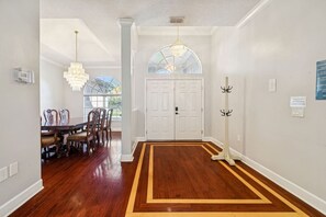 A hallway with a wooden floor, a dining area with chairs and a table, a white door, and a coat rack. - Convoy History Mansion (Tarpon Springs)