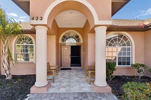 A building with a numbered address, arched windows, a central door with a 'WELCOME' sign, and two wooden chairs outside. - Convoy History Mansion (Tarpon Springs)