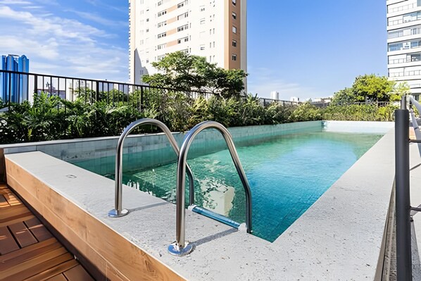 Una piscina a sfioro sul tetto, con ringhiere in metallo, circondata da vegetazione e alti edifici.