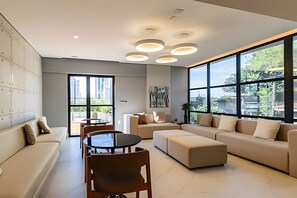 Uma sala de estar moderna com sofá, banqueta, mesa de jantar e cadeiras. Janelas amplas proporcionam luz natural e vista para o exterior.