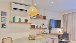 Un espace repas moderne avec une table ronde en verre, des chaises en osier, un téléviseur à écran plat fixé au mur et des étagères décoratives ornées de livres et de vases.