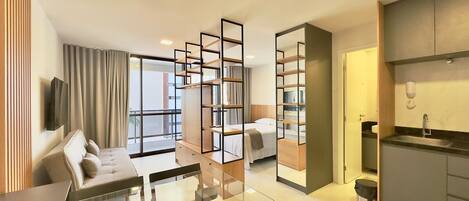 A modern hotel room with a glass dining table, a sofa, a TV, a kitchen area, and a large window.