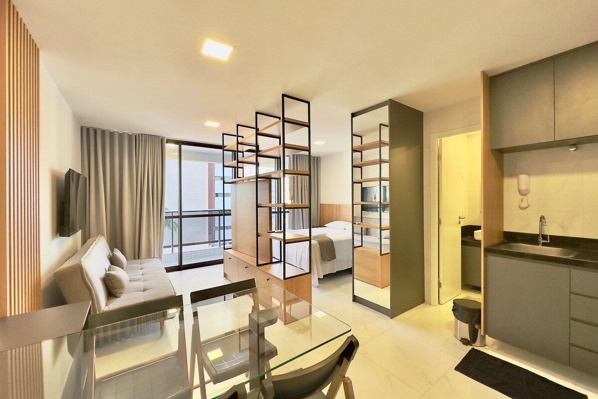 A modern hotel room with a glass dining table, a sofa, a TV, a kitchen area, and a large window.